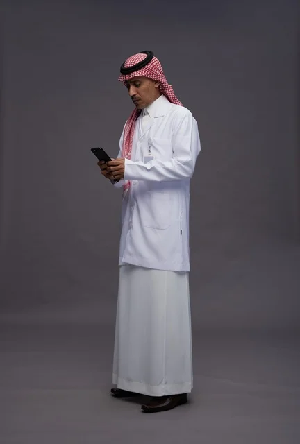 A Saudi Arabian Gulf Arab doctor wearing a thobe and shemagh with a medical coat using a mobile phone, digital health services, full body, gray background.