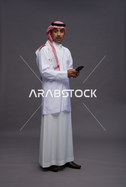 طبيب عربي سعودي خليجي يرتدي الثوب والشماغ مع المعطف الطبي يستخدم الهاتف المحمول, الخدمات الصحيه الرقميه, كامل الجسم, خلفيه رماديه