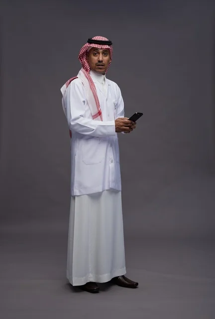 طبيب عربي سعودي خليجي يرتدي الثوب والشماغ مع المعطف الطبي يستخدم الهاتف المحمول, الخدمات الصحيه الرقميه, كامل الجسم, خلفيه رماديه