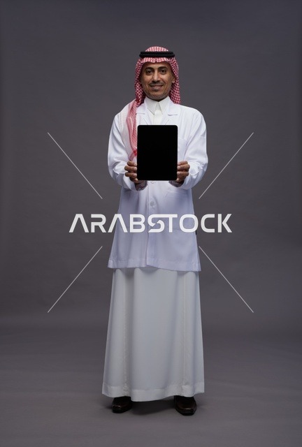طبيب عربي سعودي خليجي يرتدي الثوب والمعطف الابيض يحمل جهاز لوحي بشاشه سوداء فارغه, كامل الجسم, خلفيه رماديه