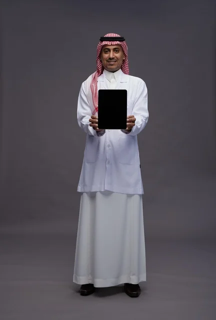 An Arab Saudi Gulf doctor wearing a thobe and a white coat holding a tablet with a blank black screen, full body, gray background.