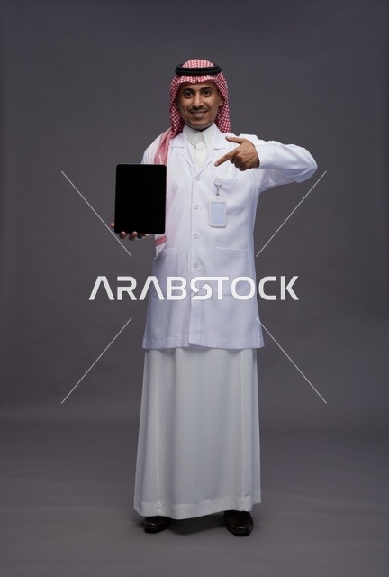 طبيب عربي سعودي خليجي يرتدي الثوب والمعطف الابيض يحمل جهاز لوحي بشاشه سوداء فارغه, كامل الجسم, خلفيه رماديه
