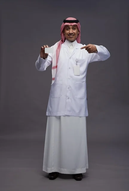 بورتريه لطبيب عربي سعودي خليجي يرتدي الثوب والمعطف الابيض يعرض بطاقة ائتمان بيضاء فارغه ويشير اليها, كرت تعريفي شخصي, الجسم بالكامل, خلفيه رماديه