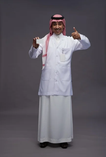بورتريه لطبيب عربي سعودي خليجي يرتدي الثوب والمعطف الابيض يعرض بطاقة ائتمان بيضاء فارغه ويشير اليها, كرت تعريفي شخصي, الجسم بالكامل, خلفيه رماديه