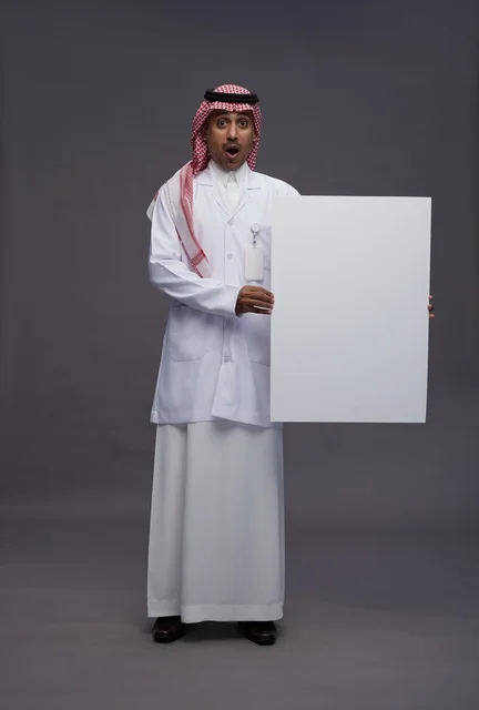 بورتريه لطبيب عربي سعودي  خليجي يرتدي المعطف الطبي يشير إلى لوح مربع ابيض فارغ , لوحه اعلانيه تسويقيه, كامل الجسم, خلفيه رماديه