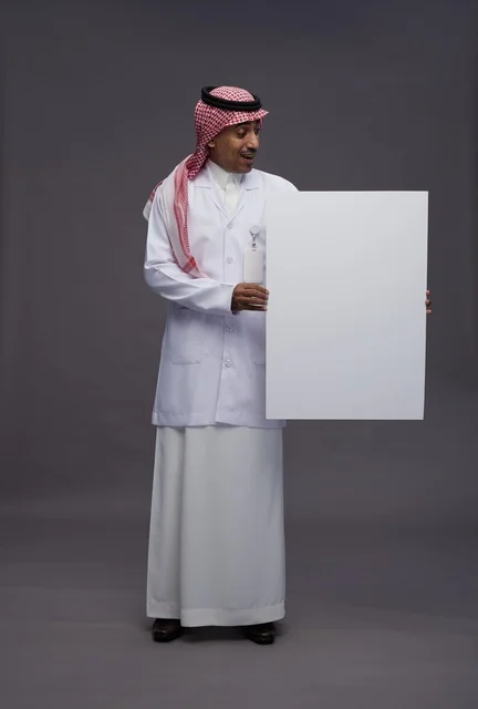 بورتريه لطبيب عربي سعودي  خليجي يرتدي المعطف الطبي يشير إلى لوح مربع ابيض فارغ , لوحه اعلانيه تسويقيه, كامل الجسم, خلفيه رماديه