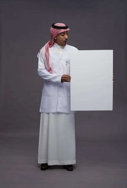 بورتريه لطبيب عربي سعودي  خليجي يرتدي المعطف الطبي يشير إلى لوح مربع ابيض فارغ , لوحه اعلانيه تسويقيه, كامل الجسم, خلفيه رماديه