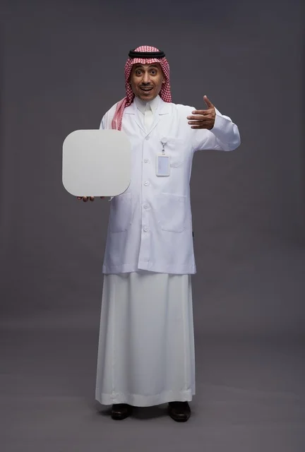 بورتريه لطبيب عربي سعودي  خليجي يرتدي المعطف الطبي يشير إلى لوح مربع ابيض فارغ , لوحه اعلانيه تسويقيه, كامل الجسم, خلفيه رماديه