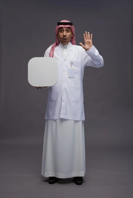 بورتريه لطبيب عربي سعودي  خليجي يرتدي المعطف الطبي يشير إلى لوح مربع ابيض فارغ , لوحه اعلانيه تسويقيه, كامل الجسم, خلفيه رماديه
