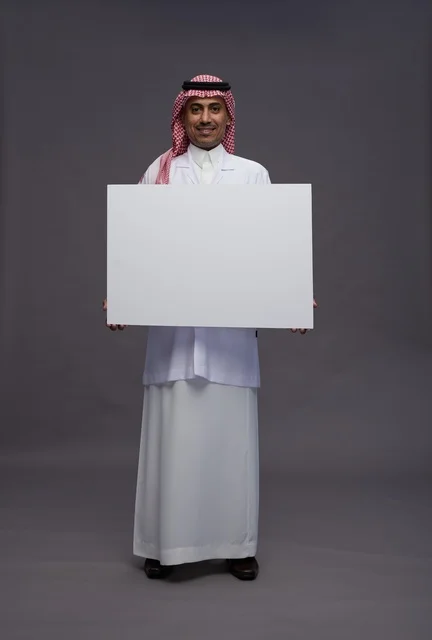 بورتريه لطبيب عربي سعودي  خليجي يرتدي المعطف الطبي يشير إلى لوح مربع ابيض فارغ , لوحه اعلانيه تسويقيه, كامل الجسم, خلفيه رماديه