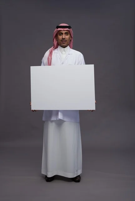 بورتريه لطبيب عربي سعودي  خليجي يرتدي المعطف الطبي يشير إلى لوح مربع ابيض فارغ , لوحه اعلانيه تسويقيه, كامل الجسم, خلفيه رماديه