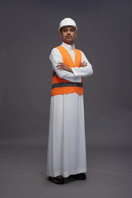 A portrait of an Arab Saudi Gulf construction engineer wearing a white thobe with a helmet and safety vest, working in the engineering sector, gray background, full body.