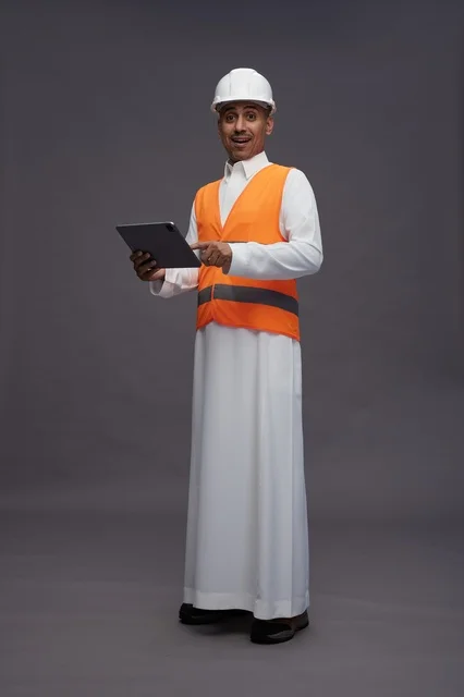 A portrait of an Arab Saudi Gulf construction engineer wearing a white thobe with a helmet and safety vest using a tablet, working in the engineering sector, gray background, full body.