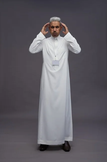 A portrait of a Saudi Arabian Gulf man wearing a white thobe, placing his hands on his head, showing tension and headache in a work environment, psychological pressure, full body, gray background.