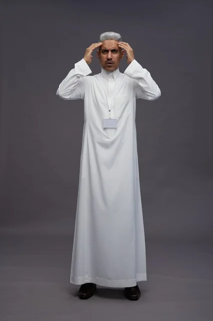 بورتريه لرجل  عربي سعودي خليجي يرتدي الثوب الابيض يضع يديه على رأسه,  التوتر والصداع في بيئة العمل, الضغط النفسي , كامل الجسم, خلفيه رماديه