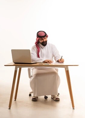 رجل سعودي يكتب على مكتب مع لابتوب بخلفية بيضاء رجل سعودي يكتب على مكتب مع لابتوب بخلفية بيضاء