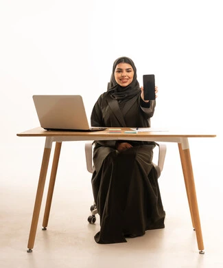 امرأة سعودية تجلس خلف مكتب عمل وتحمل هاتفاً ذكياً
