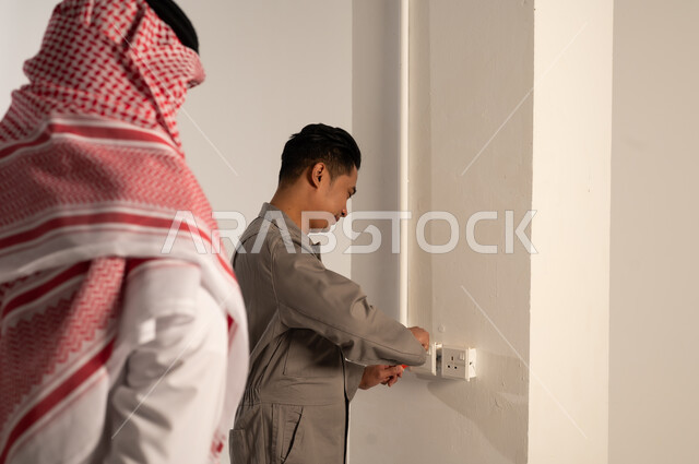 رجل سعودي يراقب فني آسيوي يصلح مقبس كهربائي