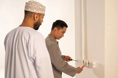 رجل عماني يراقب فني كهرباء يقوم بإصلاح المقابس