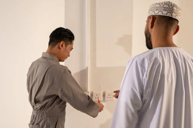 رجل عماني يراقب فني آسيوي يصلح مقبس كهرباء