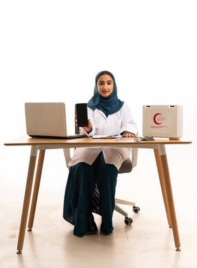 طبيبة عربية بحجاب تحمل هاتف ذكي خلف مكتب طبي