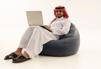 رجل سعودي بالثوب التقليدي يستخدم لابتوب على مقعد مريح