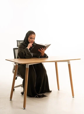 امرأة سعودية بالعباءة تقرأ كتاباً على المكتب
