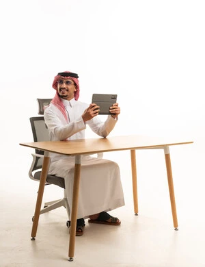 رجل سعودي يرتدي الثوب التقليدي مع جهاز لوحي على مكتب