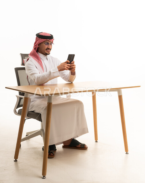 رجل سعودي بالثوب التقليدي يستخدم الجوال على المكتب