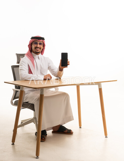 رجل سعودي بالزي التقليدي يحمل هاتف ذكي على المكتب