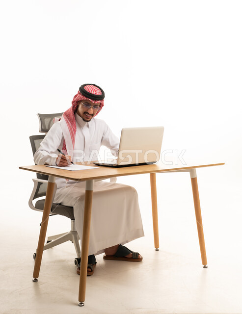 Saudi Man in Traditional Thobe Writing at Laptop Desk