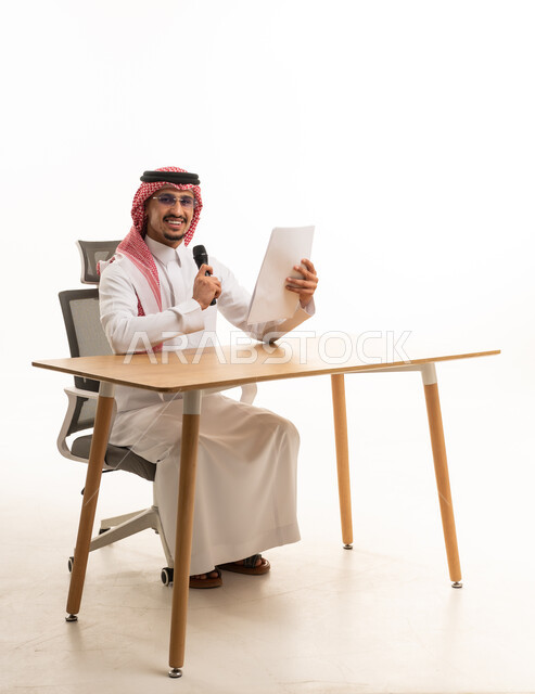 رجل أعمال سعودي خلف المكتب يمسك ميكروفون وورقة