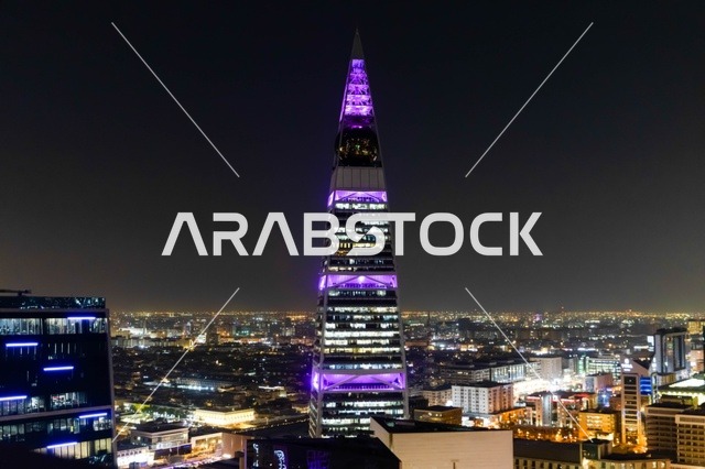 يقف برج الفيصلية شامخاً فوق أفق مدينة الرياض في المملكة العربية السعودية. تظهر العمارة الحديثة ذات الشكل الهرمي  في الليل مع إطلالة بانورامية على المدينة بالأسفل.