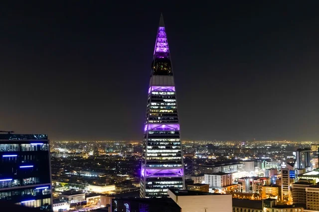 يقف برج الفيصلية شامخاً فوق أفق مدينة الرياض في المملكة العربية السعودية. تظهر العمارة الحديثة ذات الشكل الهرمي  في الليل مع إطلالة بانورامية على المدينة بالأسفل.