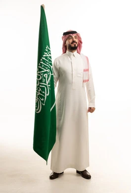 رجل سعودي بالزي التقليدي يقف مع علم السعودية