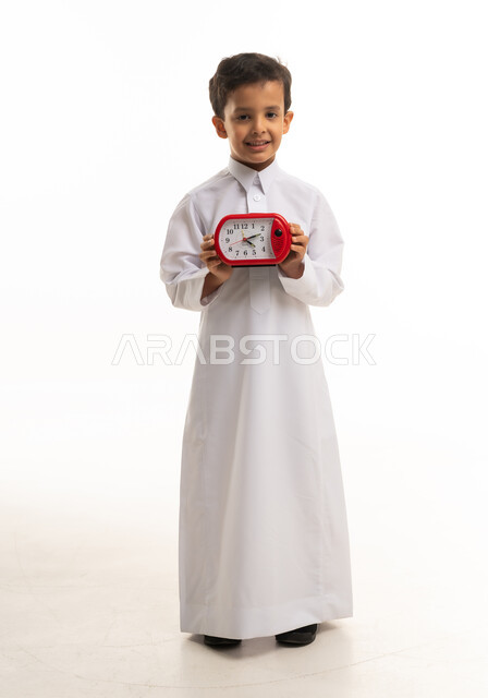 طفل سعودي بالثوب التقليدي يمسك ساعة منبه حمراء
