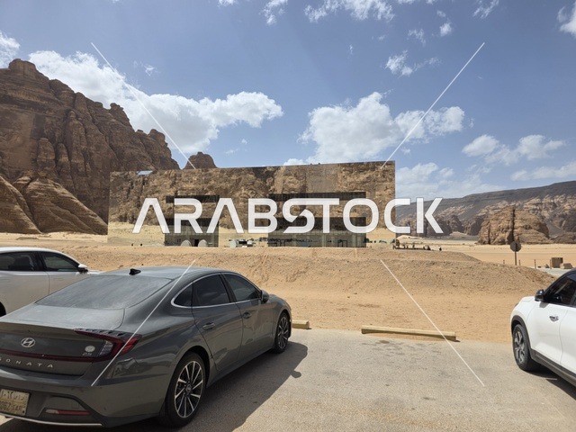 The Miraya building is located in the desert landscape of Wadi Ashar in the Al-Ula region of Saudi Arabia. The glass facade reflects the surrounding sandy mountains and the blue sky during daylight hours.
