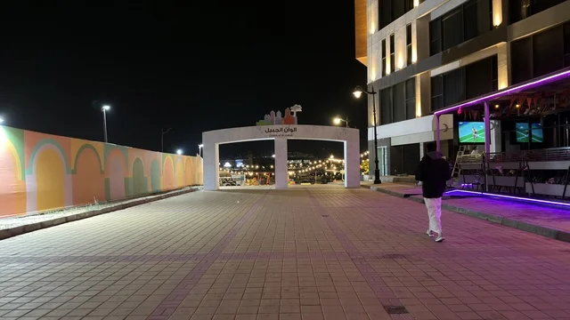 A nighttime view of the Jubail Colors event on the waterfront promenade in the Kingdom of Saudi Arabia. The decorative lights illuminate the pedestrian walkway next to modern residential buildings and palm trees.