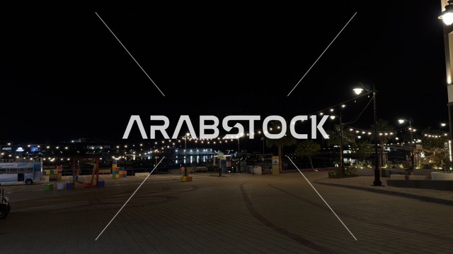 منظر ليلي لفعالية ألوان الجبيل على الممشى البحري في المملكة العربية السعودية. تضيء حبال الزينة الممشى المخصص للمشاة بجانب المباني السكنية الحديثة وأشجار النخيل.