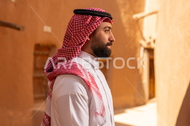 رجل سعودي بالزي التقليدي يقف أمام مباني طينية تراثية