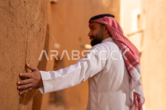 رجل سعودي يرتدي الثوب التقليدي يلمس جدار طيني في قرية تراثية