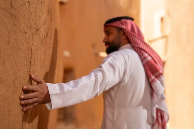 رجل سعودي يرتدي الثوب التقليدي يلمس جدار طيني في قرية تراثية