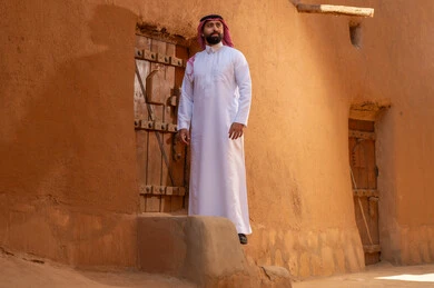 رجل سعودي يرتدي الزي التقليدي أمام منزل طيني تراثي قديم