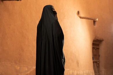 امرأة سعودية ترتدي الزي التقليدي أمام جدار طيني قديم