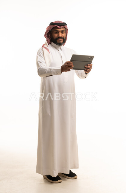 رجل سعودي واقف يحمل جهازاً لوحياً على خلفية بيضاء