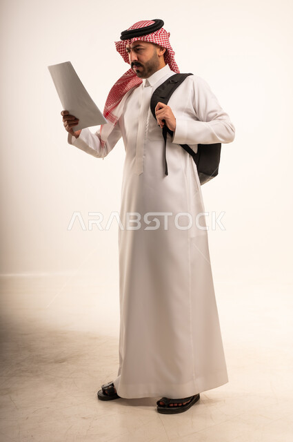 رجل سعودي بالثوب يرتدي حقيبة ظهر ويقرأ وثيقة
