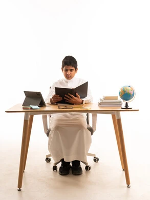 طالب سعودي بالثوب التقليدي يقرأ على مكتب دراسة في استوديو