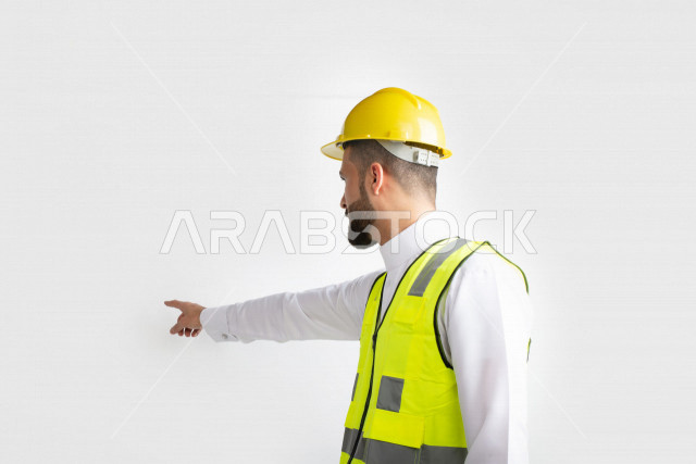 بورتريه مقرب جانبي لمهندس عربي خليجي سعودي يرتدي ستره وخوذه الحمايه، يشير باصبع السبابه الى مكان ما، الهندسه والتخطيط’ اداره المشاريع الهندسيه، خلفية بيضاء