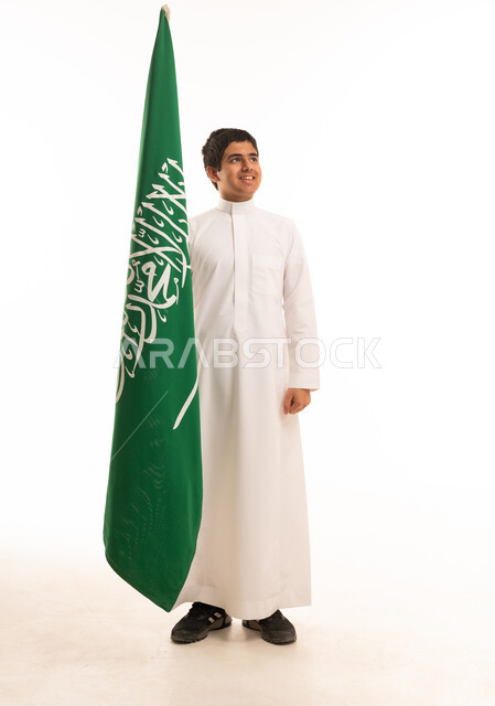 فتى سعودي يرتدي الثوب التقليدي ويقف بجانب علم المملكة