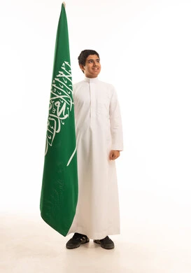فتى سعودي يرتدي الثوب التقليدي ويقف بجانب علم المملكة